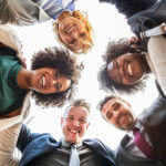 A huddle of diverse business professionals smiling and looking downward to wards the center.