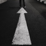 A man standing at the end of the road wearing dark pants and a bold white arrow pointing forward from behind him.