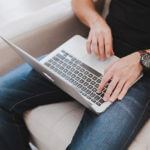 A man wearing blue jeans and a large leather watch on his left hand working on a gray laptop on his lap.