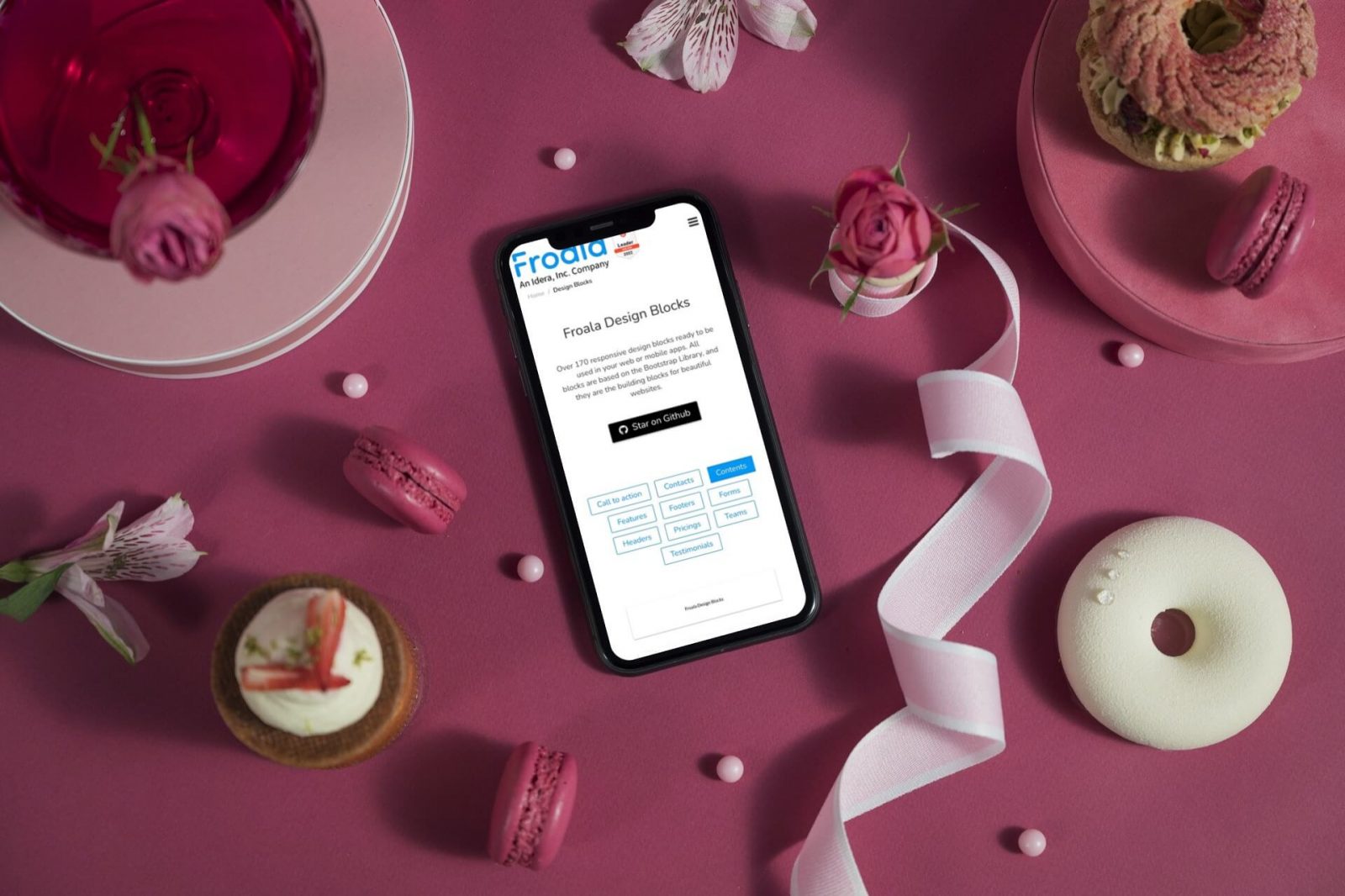 A smartphone displaying design blocks, surrounded by macarons and flowers on a pink surface.